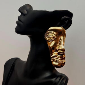 May include: A gold-colored, face-shaped earring with a smooth, reflective surface. The earring is attached to a black mannequin head, showcasing its design and form. The earring's design includes facial features like an eye, nose, and mouth.