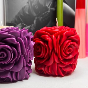 May include: Two rose-shaped candles, one red and one purple, are displayed on a white surface. The candles are made of wax and have a realistic rose design.