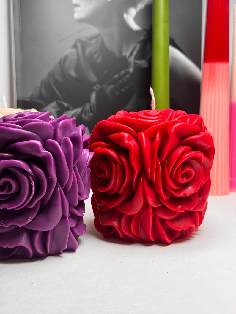 May include: Two rose-shaped candles, one red and one purple, are displayed on a white surface. The candles are made of wax and have a realistic rose design.