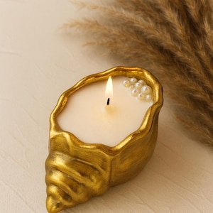 May include: A gold-colored seashell-shaped candle holder with a lit candle and small pearl accents. The candle has a white wax and a single wick. The seashell design adds a coastal and decorative touch.