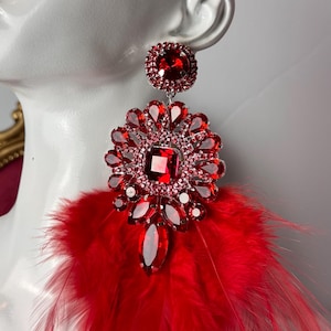 May include: A glamorous red statement earring featuring a large, square-cut red gemstone surrounded by smaller crystals. The earring has a cluster of red feathers at the bottom, adding a dramatic flair. The earring is displayed on a white mannequin head.