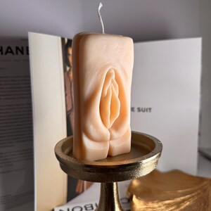 May include: A nude-colored candle shaped like a vulva, sitting on a gold metal stand. The candle is on a gold leaf-shaped surface.