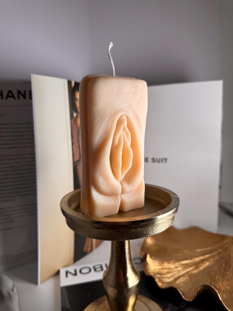 May include: A nude-colored candle shaped like a vulva, sitting on a gold metal stand. The candle is on a gold leaf-shaped surface.