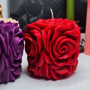May include: Two decorative candles shaped like roses. The candle on the left is purple and the candle on the right is red.
