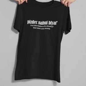 Op de afbeelding: Zwart T-shirt met witte tekst die "pizdec nahui blyat' (rus) beschrijving van een situatie die heel erg misging." zegt.