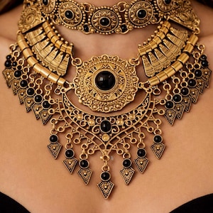 May include: Gold-toned statement necklace with intricate filigree and black accents. The necklace features multiple layers, including a choker style and a larger pendant with a central black stone. The design incorporates geometric shapes and dangling elements.