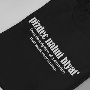 Op de afbeelding: Zwart T-shirt met witte tekst die "pizdec nahui blyat' (rus) beschrijving van een situatie die heel erg misging." zegt.