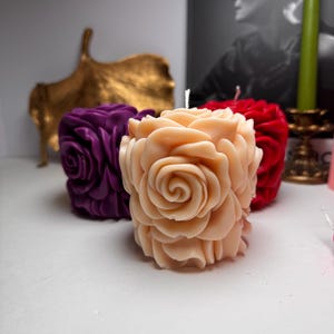 May include: Three decorative rose-shaped candles in different colours. The candles are made of wax and have a realistic rose design. The candles are arranged on a white surface.