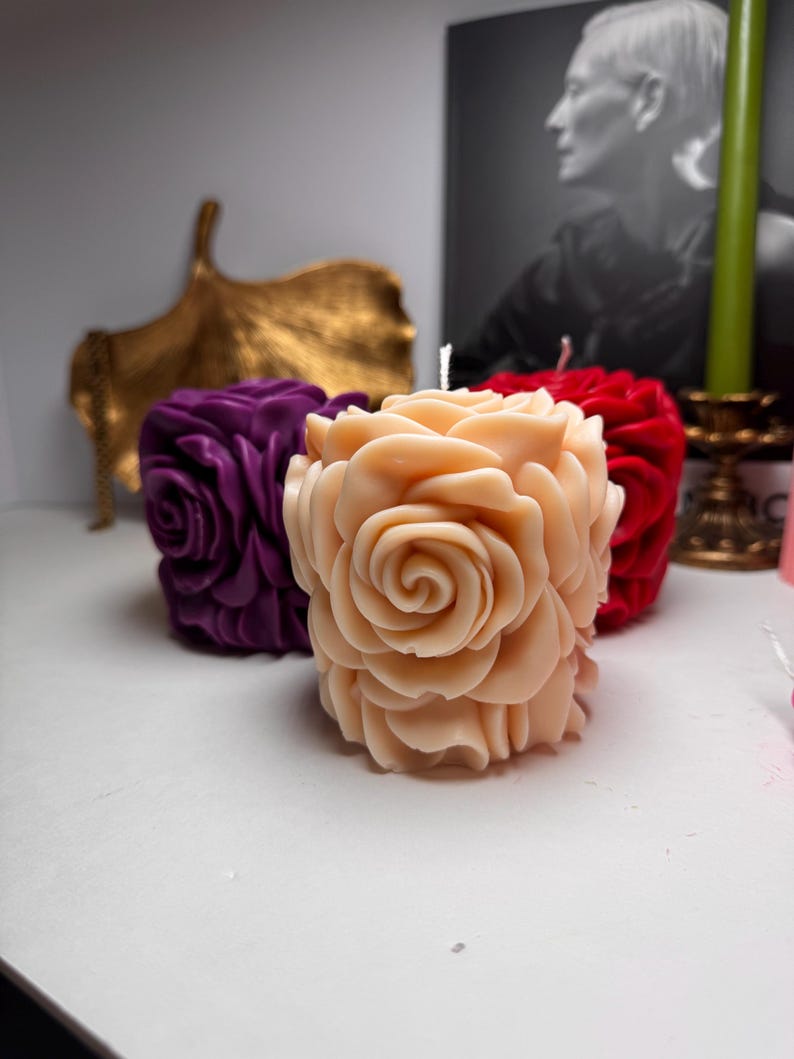 May include: Three decorative rose-shaped candles in different colours. The candles are made of wax and have a realistic rose design. The candles are arranged on a white surface.