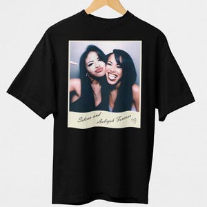 May include: Black t-shirt featuring a vintage-style photo of two women smiling. The photo has the text "Selena and Aaliyah Forever" with a heart symbol. The shirt is hanging on a wooden hanger.