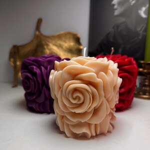 May include: Three rose-shaped candles in different colours: purple, peach, and red. The candles are arranged on a white surface.