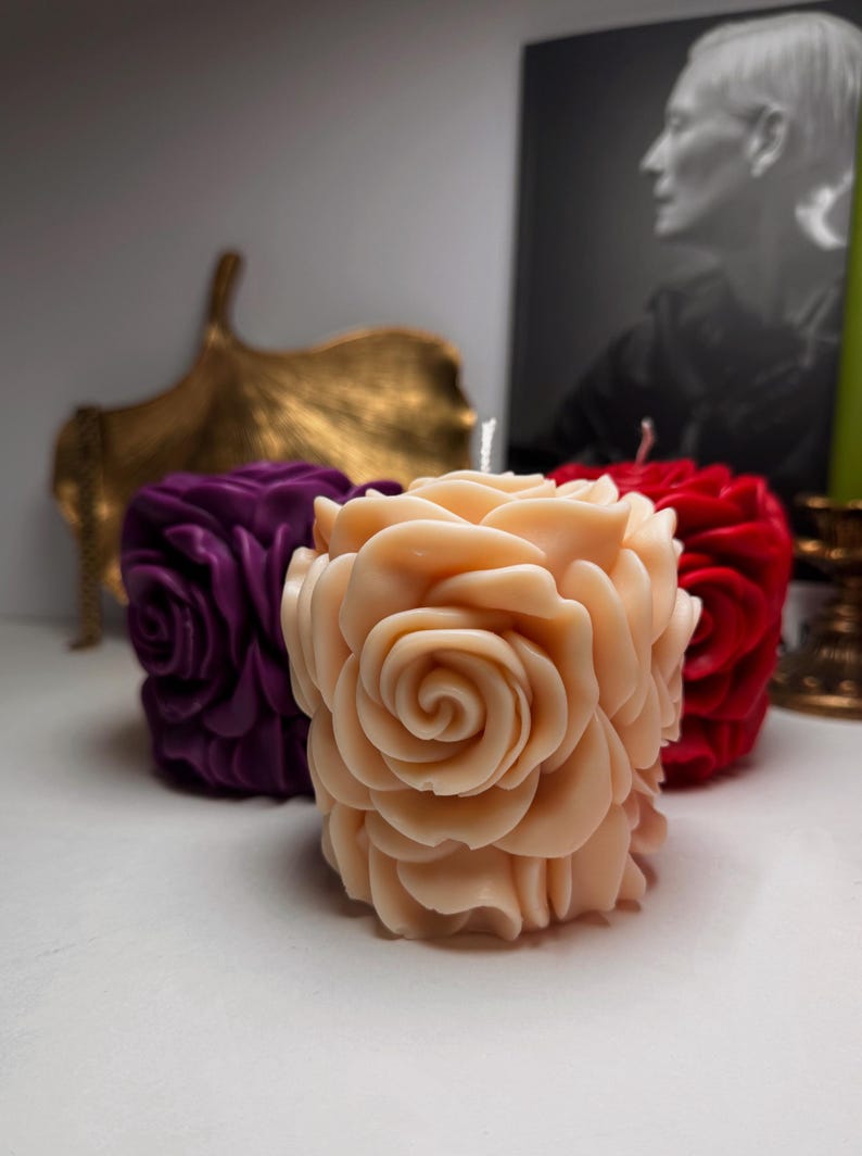 May include: Three rose-shaped candles in different colours: purple, peach, and red. The candles are arranged on a white surface.