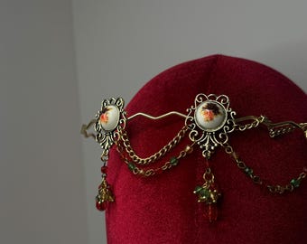 May include: A gold-toned headpiece with floral accents and dangling beads. Two oval-shaped medallions with rose illustrations are connected by delicate chains. The headpiece is displayed on a red velvet form.