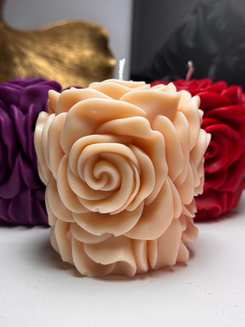 May include: A pale pink candle shaped like a rose with multiple layers of petals. The candle is on a white surface.