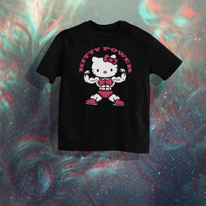 Puede incluir: Camiseta negra con un dise&ntilde;o de Hello Kitty musculosa. El dise&ntilde;o incluye el texto "KITTY POWER" en un marco circular rosa. El personaje de dibujos animados est&aacute; flexionando, con pantalones cortos rosas y un lazo.