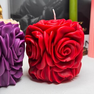 May include: Two decorative candles shaped like roses. The candle on the left is purple and the candle on the right is red.