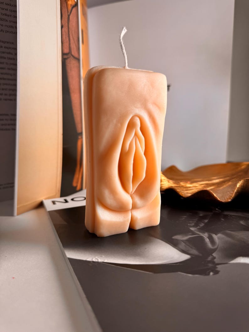 May include: A nude-colored candle shaped like a human body part. The candle has a white wick and is sitting on a black surface.