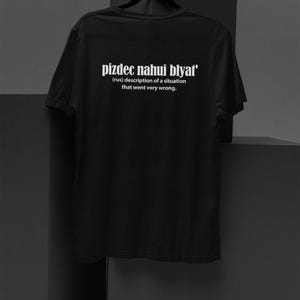Op de afbeelding: Zwart T-shirt met de tekst "pizdec nahui blyat'" in wit. De tekst is gedefinieerd als "(rus) beschrijving van een situatie die heel erg misging."