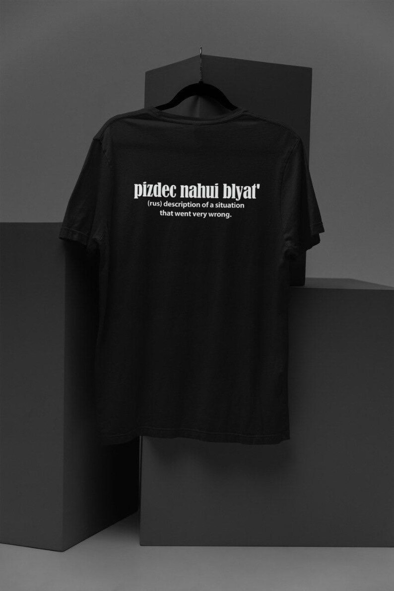 Op de afbeelding: Zwart T-shirt met de tekst "pizdec nahui blyat'" in wit. De tekst is gedefinieerd als "(rus) beschrijving van een situatie die heel erg misging."