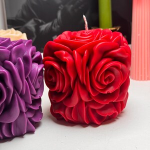 May include: Two decorative candles shaped like roses. The candle on the left is purple and the candle on the right is red.