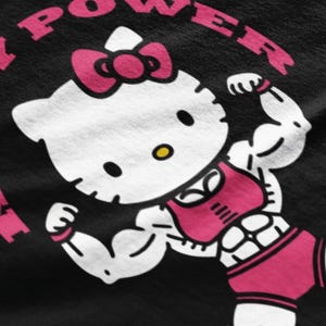 Puede incluir: Gr&aacute;fico negro con un personaje musculoso de Hello Kitty flexionando sus b&iacute;ceps. Lleva un lazo rosa, sujetador deportivo y pantalones cortos. Las palabras "KITTY POWER" est&aacute;n en rosa alrededor del personaje.