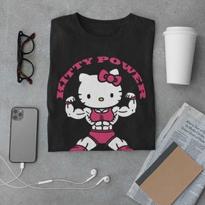 Puede incluir: Camiseta negra con un dise&ntilde;o de Hello Kitty musculosa con ropa de entrenamiento rosa, flexionando sus b&iacute;ceps. El texto "KITTY POWER" est&aacute; arqueado sobre el personaje en rosa. Otros elementos incluyen un tel&eacute;fono, gafas, auriculares y una taza de caf&eacute;.