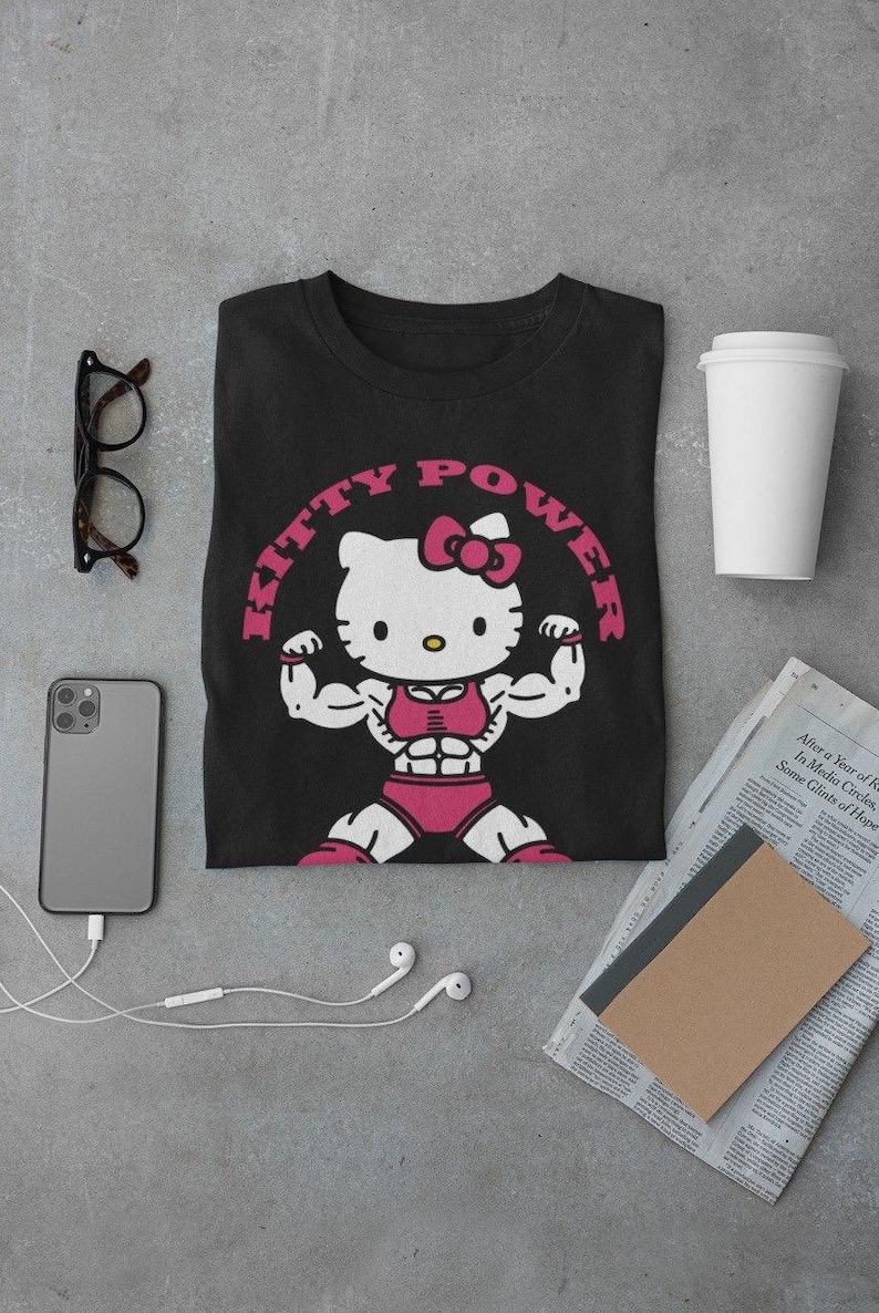 Puede incluir: Camiseta negra con un dise&ntilde;o de Hello Kitty musculosa con ropa de entrenamiento rosa, flexionando sus b&iacute;ceps. El texto "KITTY POWER" est&aacute; arqueado sobre el personaje en rosa. Otros elementos incluyen un tel&eacute;fono, gafas, auriculares y una taza de caf&eacute;.