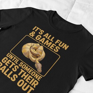 May include: Black t-shirt with a humorous graphic featuring a coiled snake inside a gold-colored rectangle. The text reads "IT'S ALL FUN & GAMES UNTIL SOMEONE GETS THEIR BALLS OUT" in gold lettering. The shirt is made of a soft material.
