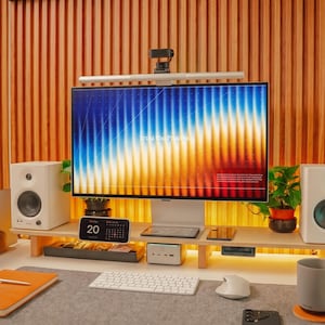 Op de afbeelding: Een computermonitor met een kleurrijk abstract scherm staat op een bureauplank van licht hout. De opstelling bevat twee witte luidsprekers, een wit toetsenbord, een computermuis, een digitale kalender, een laptopstandaard en een mok.