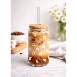 May include: A clear glass tumbler with a daisy pattern, filled with iced coffee and a clear straw. The tumbler has a bamboo lid. Coffee beans and sugar cubes are in the background. A vase of daisies is also in the background.