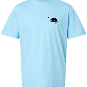 Puede incluir: Una camiseta azul claro con una silueta negra de un oso y una estrella en el pecho izquierdo. El texto "90266" está debajo del oso.