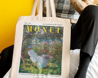 May include: Cream-coloured tote bag displaying a Monet painting, with the artist's name in yellow. The artwork shows a garden scene with a woman in a white dress. The bag has long, cream handles. The person holding the bag is wearing a black and white top.