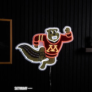 May include: A neon sign featuring a cartoon gopher mascot running, wearing a maroon sweater with a gold letter M on the chest. The gopher has brown fur, a white tooth, and is outlined in bright orange neon tubing against a dark background.