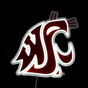 May include: A neon sign displaying the Washington State University logo, a crimson and white "WSU" Cougar head. The sign has a white outline and is set against a black background. A power cord extends from the bottom.