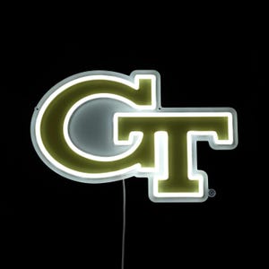 May include: A neon sign displaying the letters "GT" in gold and white. The letters are illuminated with bright white neon against a black backdrop. A power cable extends from the base of the sign.