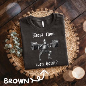 May include: Brown t-shirt with the text "Dost thou even hoist?" above a graphic of a knight lifting weights. The shirt is displayed on a woven, circular placemat with a wooden background.