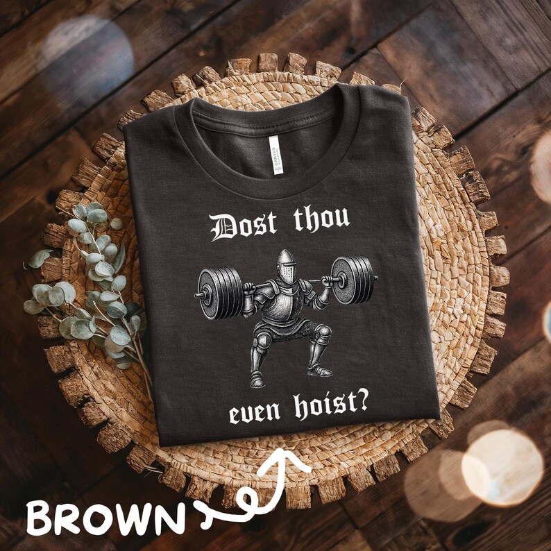 May include: Brown t-shirt with the text "Dost thou even hoist?" above a graphic of a knight lifting weights. The shirt is displayed on a woven, circular placemat with a wooden background.
