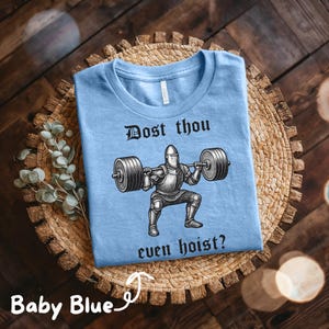 May include: Baby blue t-shirt with the text "Dost thou even hoist?" above a graphic of a knight lifting weights. The shirt is displayed on a woven, circular placemat.