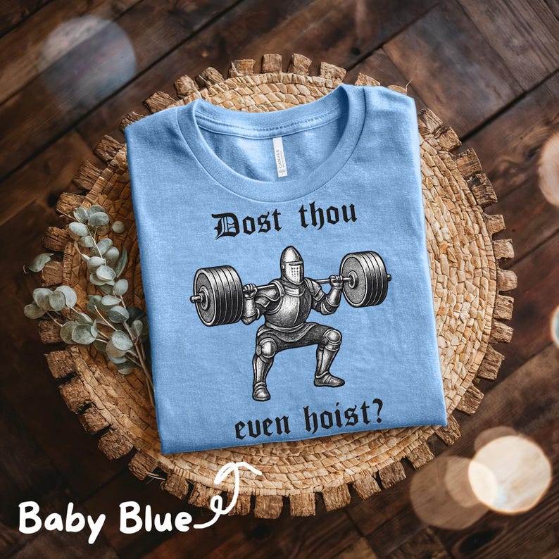May include: Baby blue t-shirt with the text "Dost thou even hoist?" above a graphic of a knight lifting weights. The shirt is displayed on a woven, circular placemat.