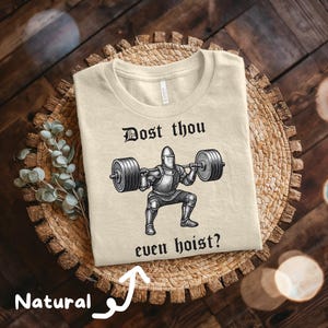 May include: Beige t-shirt with a graphic of a knight lifting a barbell, with the text "Dost thou even hoist?" in black. The shirt is made of a soft, natural-colored fabric.