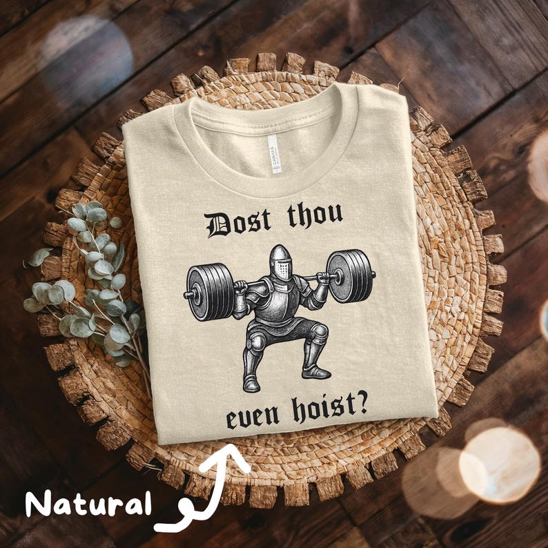 May include: Beige t-shirt with a graphic of a knight lifting a barbell, with the text "Dost thou even hoist?" in black. The shirt is made of a soft, natural-colored fabric.