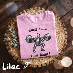 May include: Lilac-colored t-shirt with the text "Dost thou even hoist?" and a graphic of a knight lifting weights. The shirt is displayed on a woven basket with a sprig of greenery.