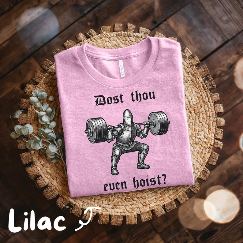 May include: Lilac-colored t-shirt with the text "Dost thou even hoist?" and a graphic of a knight lifting weights. The shirt is displayed on a woven basket with a sprig of greenery.