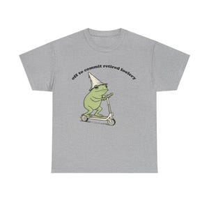 May include: A heather gray t-shirt featuring a graphic of a green frog wearing a pointed white hat and riding a kick scooter. Above the frog, the text reads, "off to commit retired foolery" in a black, curved, sans-serif font.
