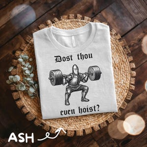 May include: A light gray t-shirt with a graphic of a knight lifting weights, with the text "Dost thou even hoist?" in black. The shirt is on a woven basket, with a wooden background.