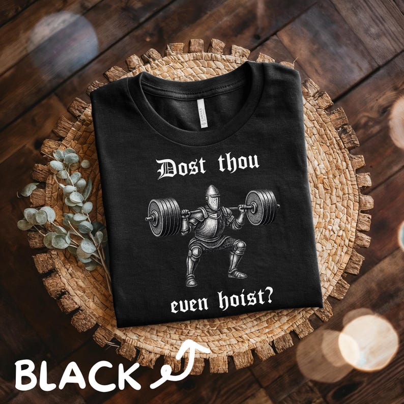 May include: Black t-shirt featuring a weightlifting knight graphic with the text "Dost thou even hoist?" in white. The shirt is displayed on a woven placemat with a wooden background. The word "BLACK" is written in white with an arrow.