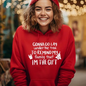 May include: A red hooded sweatshirt with the text "Gonna go lay under the tree to remind my family that I'm the gift" in white. The sweatshirt has a drawstring hood and a front pocket. A Christmas tree and gift box are also printed on the sweatshirt.
