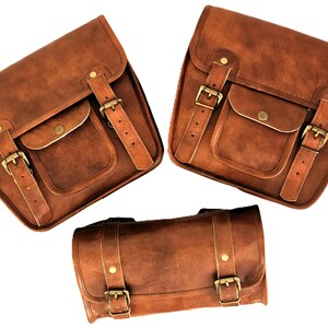 May include: Three brown leather motorcycle saddlebags with buckles and straps. The two larger bags have a flap with a buckle closure and a smaller pocket on the front. The smaller bag is cylindrical with two buckles.