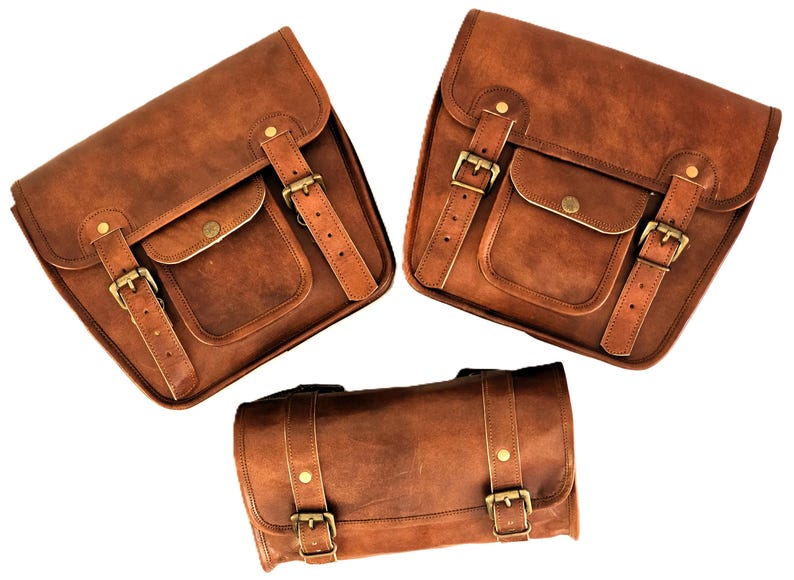 May include: Three brown leather motorcycle saddlebags with buckles and straps. The two larger bags have a flap with a buckle closure and a smaller pocket on the front. The smaller bag is cylindrical with two buckles.