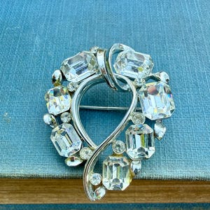 May include: A silver tone brooch with a heart-shaped design, featuring a cluster of clear, faceted rhinestones.